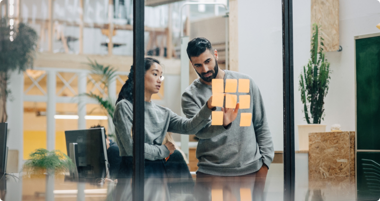 A man and woman look at post-it notes on a glass wall